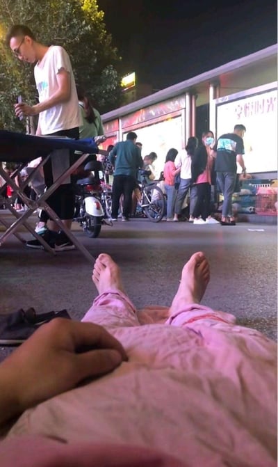 这是第一人称视角的生活随拍，拍摄者躺在夜晚的街头地面上，露出双脚和一只手，姿态十分放松；背景是热闹的市井夜市场景，左侧有站在折叠桌旁的摆摊男子、停放的电动车，远处还有一群人聚集在写有“宠喵时光”的店铺前，看起来像是在围观售卖的宠物，整体氛围充满了市井夜晚的悠闲感，拍摄者以躺平的方式在热闹的夜市中享受片刻的松弛。