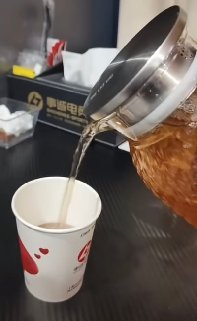办公室场景中，一只透明玻璃茶壶正在将琥珀色的茶水倒入白色纸杯中。茶壶顶部有银色不锈钢滤网，水流呈现清晰的弧线。背景可见黑色纸巾盒（印有“事诚电子”字样）、透明方糖罐和其他办公用品，营造出典型的办公环境氛围。画面聚焦于倒茶动作的瞬间，展现日常工作中的一个平静场景。