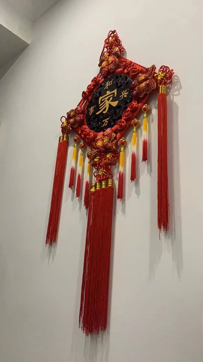 图片展示了一个悬挂在白色墙面上的传统中国结装饰。中国结主体为菱形，以大红色为主色调，辅以金色刺绣花纹，中央是黑色圆形面板，上面用金色字体书写着“家和万事兴”五个汉字，寓意家庭和睦则事事顺利。中国结边缘装饰有金色的鱼形图案，象征吉祥有余，下方垂挂着多组红色流苏，部分流苏带有黄白渐变的配色，整体造型喜庆且富有传统韵味。