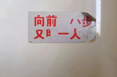 图片展示了一张贴在白色平面上的银色贴纸，贴纸表面有红色的中文文字。贴纸右上角有明显的撕裂痕迹，边缘部分破损，导致部分文字可能不完整。可见的文字内容为“向前 八步 又 一人”，文字排列较为紧凑，整体给人一种因长时间使用或意外损坏而显得陈旧的感觉。