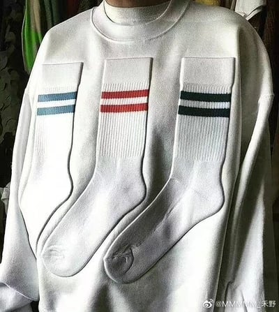 图片中展示了一件白色圆领卫衣，胸前位置整齐排列着三双白色袜子。每双袜子的袜口处都有不同颜色的横向条纹：左侧袜子为蓝色条纹，中间为红色条纹，右侧则是绿色与黑色条纹。背景中可以看到一些悬挂的衣物，整体画面聚焦于这件设计独特的卫衣，给人一种创意穿搭的视觉效果。