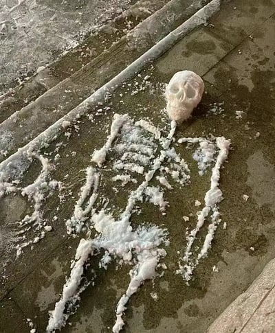 这张图片展示了在台阶上用雪堆积拼成的人体骨架造型，头骨部分更为立体，身体部分则用雪简单勾勒出肋骨和四肢。看起来好像是一个‘雪人’死亡后只剩下骨头，充满黑色幽默和恶搞氛围。这种造型一般是在冬天有人利用地上的雪随手捏造，小小恶趣味让人忍俊不禁。

没有明显的谐音梗或双关，但“雪人做成骨架”本身自带视觉上的反差萌和猎奇效果。