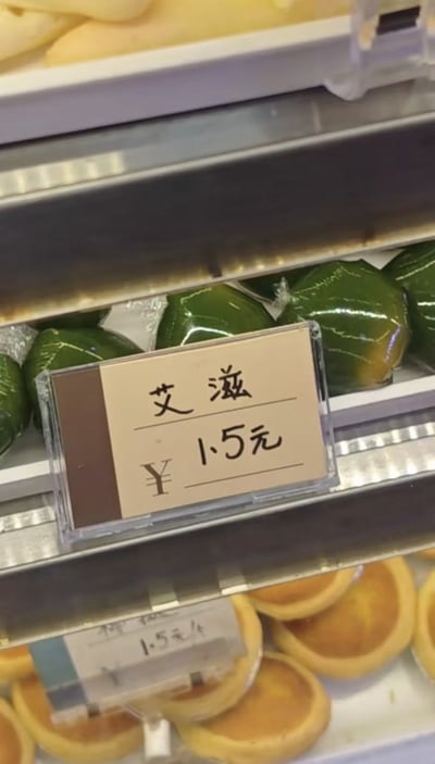 这是一张线下店铺点心货架的实拍照片，中层货架摆放着数个透明塑料包装的绿色青团，青团前方放置透明框手写价格牌，标注商品名称为“艾滋”，单价1.5元，下方货架摆放多个空蛋挞挞底，上方货架还有其他浅黄色面点；原本青团应为“艾草青团”，此处被错写为“艾滋”，谐音容易引发歧义联想，形成了搞笑的错误。