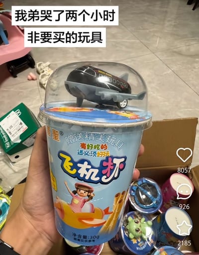 这是一张网友分享的实拍内容，图中一只手拿着名为“飞机杯”的儿童零食杯，杯盖透明罩内放置着黑色小飞机玩具，杯身标注这是附赠玩具的零食产品，上方配文“我弟哭了两个小时 非要买的玩具”。由于“飞机杯”同时是成人情趣用品的通用名称，搭配弟弟哭闹非要购买的前置描述，形成了强烈的荒诞反差笑点。背景可见居家地面、其他同款零食杯、纸箱等物品，右侧带有短视频平台的点赞、评论、收藏数据标识。