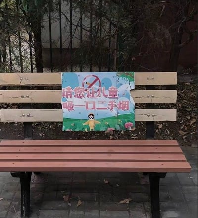 这是一张户外休闲长椅的实拍照片，长椅的靠背上贴着一张公益标语牌。原本的标语应该是“请您不要让儿童吸一口二手烟”，但标语牌的“不要”字样被损坏缺失，导致标语变成了完全相反的“请您让儿童吸一口二手烟”，形成了极具反差感的乌龙效果。背景是户外的树木与地面，环境看起来是公园或者社区的休闲区域，整体场景真实且充满意外的搞笑感。