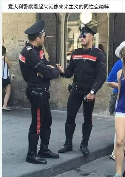 这张图片拍摄于意大利街头，画面中心是两名身着黑红配色紧身制服的意大利警察，他们站在具有复古石质建筑风格的街道边。左侧警察双臂交叉站立，侧脸对着同伴，戴着墨镜和带帽檐的警帽；右侧警察同样戴着墨镜和警帽，抬手做出动作，似乎正在和左侧警察交流。两人的制服搭配高筒靴，造型硬朗利落。图片顶部配有中文调侃文字“意大利警察看起来就像未来主义的同性恋纳粹”，这只是网友对其制服外观风格的趣味评价，无实际政治指向。画面中还能看到几名路人，背景是带有拱形门窗的欧式建筑，充满异国街头氛围。