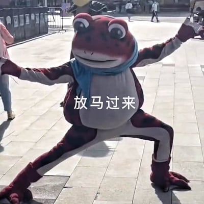 图片中是一个穿着红色青蛙吉祥物服装的人，戴着蓝色围巾，在户外广场上摆出双臂张开、双腿分开的姿势。人物面带微笑，姿态显得自信且充满活力。图片中央有白色文字“放马过来”，传达出一种挑战或欢迎的意味。背景中可以看到模糊的行人和广场设施，显示这是在一个公共场所拍摄的真实场景。
