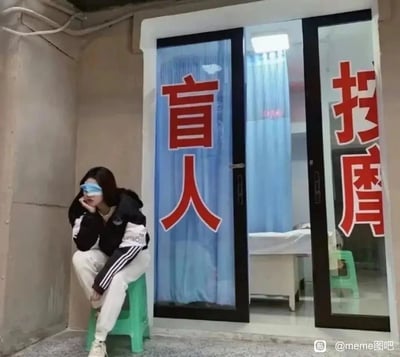 图片呈现了一处室内角落场景，一位身着黑白拼色连帽外套、白色侧边带黑条纹运动裤的女生，佩戴着蓝色眼罩，坐在绿色小矮凳上，手肘搭在弯曲的膝盖上，一只手托着脸颊，姿态流露出惆怅无聊的感觉。她身后是盲人按摩店的玻璃推拉门，门上贴着醒目的红色“盲人”“按摩”字样，透过玻璃能看到店内的按摩床与蓝色帘子，地面为水泥质感，整体环境简单朴素。这张图通过女生的眼罩和身后的盲人按摩店形成趣味呼应，制造出她就是店内盲人按摩师的错觉，属于玩梗类搞笑图片。