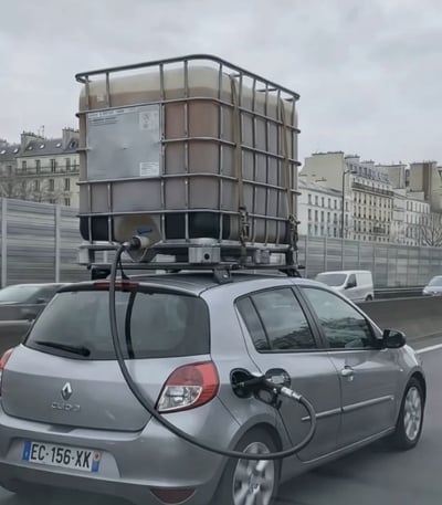 这张实拍照片拍摄于城市高架道路上，主体为一辆悬挂法国车牌的银色雷诺Clio3小轿车正在行驶，车辆的车顶行李架上用绑带固定了一个带外部金属防护框架的大容量工业塑料储液吨桶，吨桶底部的出口接出黑色油管，一路延伸到车辆侧面的加油口内，相当于私自为车辆加装了远超常规容量的额外油箱，背景可见典型的欧洲城市联排建筑、道路隔音护栏以及其他行驶中的社会车辆，天空阴沉。