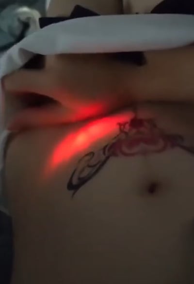 图片展示了一个人的腹部区域特写，皮肤上有一个黑色线条构成的纹身图案，图案设计包含类似花朵和卷曲线条的元素。纹身区域被一个红色光源照亮，形成明显的红色光斑效果。光源似乎来自手持设备，光线集中照射在纹身中心位置，使该区域与周围皮肤形成鲜明对比。