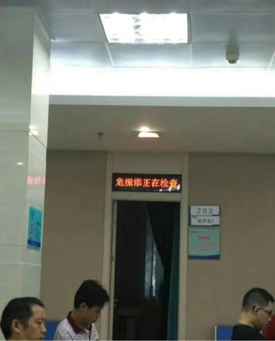 这是一张在医院走廊门口拍摄的照片，门上方的LED屏显示“危振沈正在检查”。这句话本意为“危重症正在检查”，但由于LED板显示的字体间隔或分组，读作“危振沈正在检查”，读起来像是一个人的姓名（危振沈）正在检查，产生了有趣的双关或谐音梗，令人忍俊不禁。这种类似“奇葩中文分隔”的效果，经常在网络梗图中被用来调侃生活场景的荒谬，非常适合网络传播。

本图虽然拍摄于真实医院，但因为屏幕显示的原因带有幽默感，无明显政治或敏感内容。