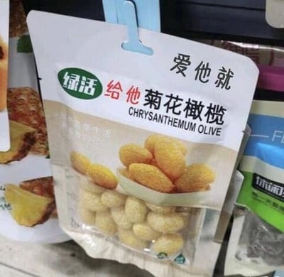 图片展示了一款名为“绿活 给他菊花橄榄”的零食包装，包装顶部印有醒目的文案“爱他就给他菊花橄榄”，下方是产品名称及英文“CHRYSANTHEMUM OLIVE”，中间配有橄榄实物图片。背景中可见其他零食包装，推测拍摄于超市货架。整体画面清晰，聚焦于该款零食的包装设计，文案表述较为独特。