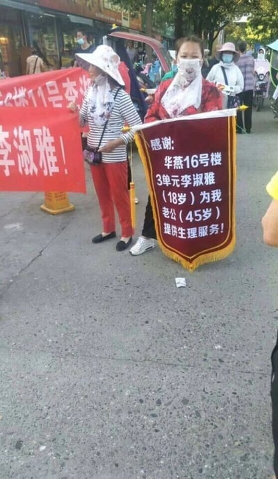 这是一张户外街道场景的实拍照片，画面核心是两位女士：左侧女士头戴宽檐遮阳帽，身着黑白条纹上衣与红色长裤，双手举着一条红色横幅，横幅上可见白色文字“2栋11号李”以及“李淑雅！”；右侧女士佩戴口罩，身穿红色花纹上衣，身前悬挂着一面红底黄字的锦旗，锦旗上的文字内容极具争议性，写着“感谢:华燕16号楼3单元李淑雅（18岁）为我老公（45岁）提供生理服务！”。背景中能看到街边的商铺、树木以及其他路过的行人，整体场景是公开的城市街道，事件性质属于引发争议的公开举牌行为，文字内容暗示了违背公序良俗的特殊关系。