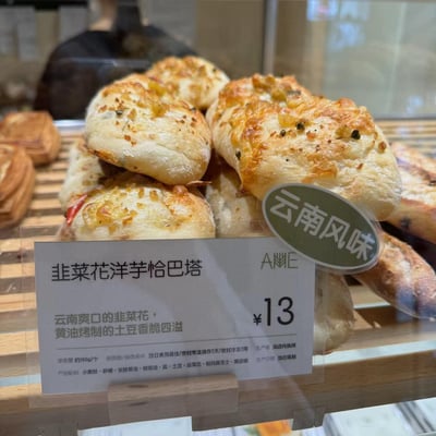 面包店展示柜中堆叠着金黄诱人的韭菜花洋芋恰巴塔，表面撒有翠绿的韭菜花碎和融化的芝士，呈现出烘烤后的焦香色泽。面包旁立着透明标牌，标注着产品名称、特色描述“云南爽口的韭菜花，黄油烤制的土豆香脆四溢”以及13元的价格。背景中隐约可见其他烘焙产品，整体画面突出了这款融合云南风味的西式面包。