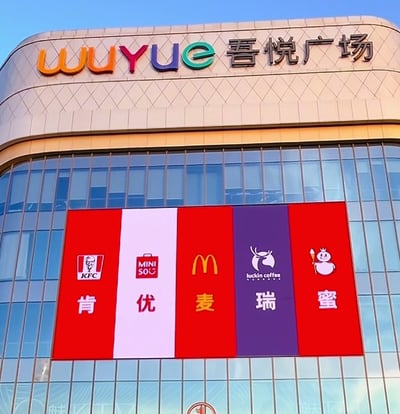 这张图片拍摄于吾悦广场外墙，画面上主要展示了五大热门品牌的Logo与对应汉字，依次是KFC（肯）、名创优品（优）、麦当劳（麦）、瑞幸咖啡（瑞）、蜜雪冰城（蜜），组合后谐音为“肯优麦瑞蜜”。这种拼贴方式利用了品牌名称的发音和中文字组成，形成幽默有趣的商圈谐音梗，画风现代感十足，很适合用来调侃品牌扎堆现象。

图片属于纯趣味拼贴，没有明显暗示成分，仅为商业文化梗。