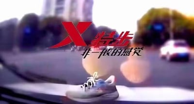 这是一张特步品牌的广告视频截图，画面前景是一只放置在汽车仪表台上的特步运动鞋，鞋款为浅色系搭配蓝红细节设计；背景是虚化的城市道路场景，能看到行驶的车辆与道路设施，营造出动态的氛围。画面上方配有醒目的红色“X特步”品牌标识，下方是黑色的品牌经典标语“非一般的感觉”，通过将运动鞋置于车内的创意场景，结合背景的道路动态感，呼应品牌标语，传递出该运动鞋能带来畅快、与众不同的运动体验的宣传核心。