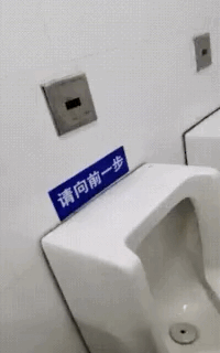 这是一张竖屏的原始拍摄动态图，展示了男厕所内的场景。白色墙面上的小便池正上方贴着一块蓝色的提示牌，上面写着“请向前一步”，但由于标语的安装位置失误，形成了视觉错位效果，原本提示使用者向前凑近便池的文明标语，看起来像是要使用者凑近这个牌子，和原本的意图完全偏离，制造出荒诞搞笑的反差感。