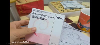 这是B站视频的截图，画面中一只手拿着Debian GNU/Linux的盒装安装媒介，包装上印有「请仔细阅读安装说明并在管理员指导下使用，谨慎用于服务器安装」的提示语与产品名称。背景桌面摆放着KFC包装袋、一盒中国象棋、带图案的咖啡杯以及画有线条的纸张，呈现出日常桌面的场景，似乎是在进行该系统安装介质的开箱展示。