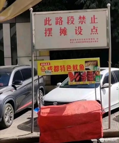 这张图片呈现了极具讽刺感的现实场景：画面上方是由攀枝花市东区城市管理局、东区人民政府炳草岗街道办事处设立的白色告示牌，上面用红色字体明确写着“此路段禁止摆摊设点”；而在告示牌的金属支架下方，直接设置了一个“成都特色鱿鱼”的小吃摊，摊位的黄色宣传牌上印有鱿鱼产品图片和收款二维码，摊位主体被红色布罩覆盖。背景中可以看到停放的两辆汽车，整个场景形成了官方禁令与实际摆摊行为的强烈反差，充满了荒诞的讽刺意味。