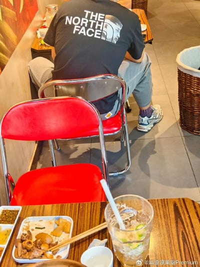 这是在一家餐馆内的日常用餐场景，前景摆放着食用过半的餐品与一杯带吸管的饮品。画面中一位背对镜头的顾客穿着一件恶搞THE NORTH FACE品牌的定制T恤——将品牌标志性的雪山标识替换为一张人脸，与品牌文字「THE NORTH FACE」组合，形成反差感拉满的趣味效果。周围是红色折叠椅与餐馆的地面环境，整体氛围生活化，恶搞T恤是画面的核心趣味亮点。