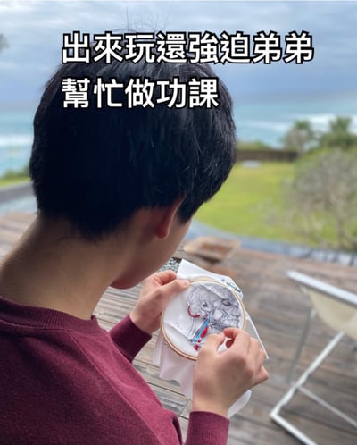 图片中一名身穿酒红色毛衣的男子站在木质甲板上，背对着镜头，手里拿着一个刺绣绷子，正在绣一幅精细的人体头部解剖图。背景是开阔的海景和绿色草地，天空有些阴沉。图片上方配有文字“出来玩还强迫弟弟帮忙做功课”，形成一种幽默的反差感——在度假般的环境中却在进行类似功课的精细刺绣工作。