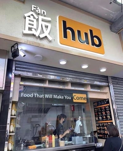 这是一张街头餐饮门店的实拍图，门店招牌采用中英结合的设计，上方是白色立体字“饭 Fàn”搭配黄色底的黑色“hub”字样，构成店名“饭hub”。门店玻璃上贴有标语“Food That Will Make You Come”，其中“Come”被单独放在黄色方框内，玩了双关梗：字面意思是“这里的食物会吸引你前来”，同时“Come”和店名的“hub”形成趣味呼应，结合中文“饭”，也能让人联想到“饭来”的谐音趣味。画面中店内有一位戴口罩的工作人员，店外有一位顾客正在看菜单，整体是日常的街头餐饮门店场景。