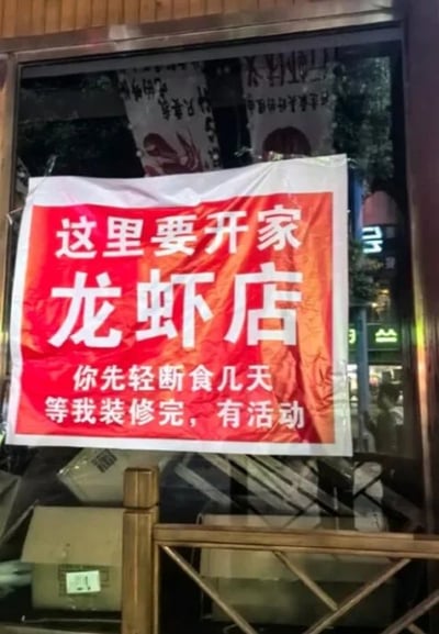 这是一张实拍的线下商铺照片，拍摄时间为夜晚。待装修的龙虾店玻璃上张贴了一张红底白字的横幅，横幅内容以调侃的语气告知路人此处即将开设龙虾店，还开玩笑让大家先轻断食几天留好胃口，等待店铺装修完毕后参与开业活动，风格接地气又有网感，玻璃内部可以看到装修相关的纸箱，玻璃外能看到夜晚街道的灯光倒影。