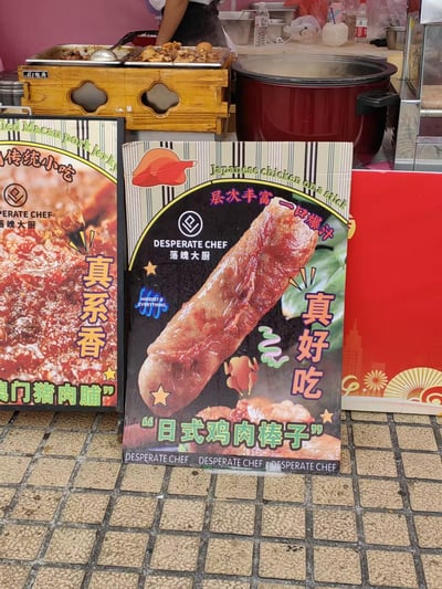 街头小吃摊的特写照片，展示了两个主要广告牌和烹饪场景。前景中央是"DESPERATE CHEF 落魄大厨"的日式鸡肉棒子广告，画面上有一根油亮的鸡肉棒特写，配文"层次丰富 一口爆汁 真好吃"。左侧部分可见另一个广告牌，宣传"澳门猪肉脯"，写着"真系香"。背景中能看到冒着热气的烹饪设备和食材，一位穿着围裙的摊主正在准备食物，营造出热闹的街头美食氛围。