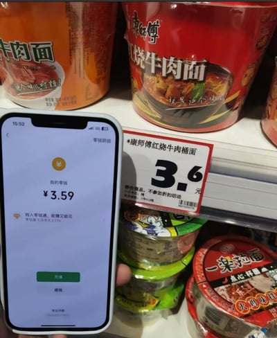 这是一张在超市货架前拍摄的实拍图，画面里手机显示零钱余额为3.59元，而旁边货架上的康师傅红烧牛肉桶面售价3.6元，只差1分钱就能购买，精准呈现了日常里没钱的窘迫与尴尬。图中没有双关、谐音梗或暗示内容。