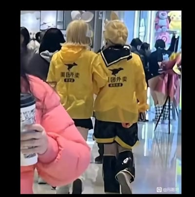 两名穿着美团外卖黄色制服的coser在漫展现场行走，制服背后印有美团外卖标志和文字。两人都戴着黄色假发和动物耳朵，周围有许多其他游客和动漫周边展示，营造出一种创意混搭的有趣氛围。
