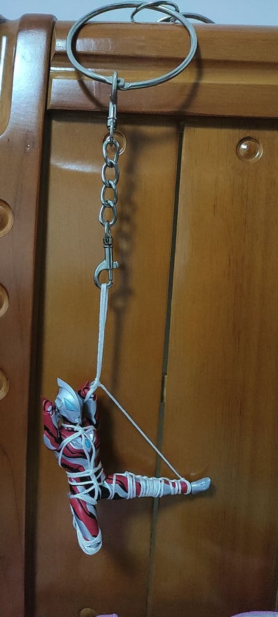 图片展示了一个红银配色的奥特曼钥匙扣，其身体和四肢被白色绳子紧紧捆绑，绳子形成环状连接到钥匙扣的金属链条。钥匙扣通过圆形金属环悬挂在带有圆形旋钮的木质柜子顶部，奥特曼经典的严肃表情与被束缚的滑稽姿态形成反差萌。画面无任何文字内容，整体呈现出一种手工改造的趣味搞笑效果。