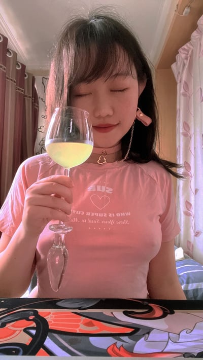 图中是一位年轻女生在室内的居家场景。她穿着一件粉色T恤，T恤上印有白色文字“SUE WHO IS SWEETER THAN... YOUR SOUL IS SWEET AS...”，脖子上戴着一条带有心形吊坠的项链，耳朵上佩戴着粉色耳饰。她右手手持一个装有淡黄色液体的高脚杯，眼睛微闭，嘴角上扬，面带微笑，神情放松惬意。背景中可以看到带有花纹的粉色窗帘和部分床铺，整体环境温馨舒适，光线柔和，营造出一种悠闲自在的日常氛围。