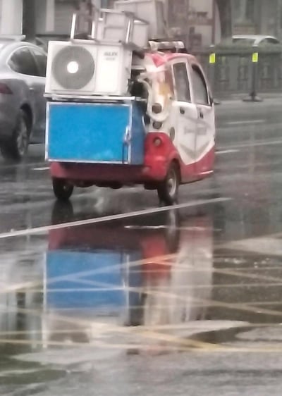 雨后积水的城市路面上，一辆红白配色的三轮老年代步车被民间硬核改装，车尾加装了完整的奥克斯空调外机与蓝色箱体，车辆正在湿滑路面行驶，积水清晰倒映出车辆的轮廓，充满生活脑洞感，展现了接地气的民间改装趣味场景。