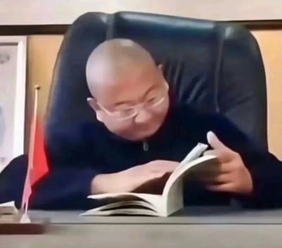 这是一张经过P图改编的静态趣味图，画面呈现出视觉错位的效果，一位光头戴细框眼镜的男士头部和趴在桌上的身体被合成在一起，男士身着深色上衣，坐在黑色办公老板椅上，双手捧着一本翻开的书，低头专注地看着书页，脸上还带着笑意，仿佛看得十分投入。办公桌的左侧摆放着一面小型的红旗摆件，整体场景模拟办公室环境，营造出一种假装认真阅读的搞笑氛围，常被用作调侃摸鱼或假装努力的搞笑表情包。