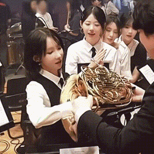这是一段记录校园乐团日常的动态GIF，画面中一位穿着乐团制服的女生正双手接过他人递来的圆号，圆号复杂的金色管体在这个角度下看起来像是一堆面条，女生脸上露出惊讶又好奇的表情，周围的同学则笑着看向她，整个场景轻松欢乐，充满了校园生活的趣味性。