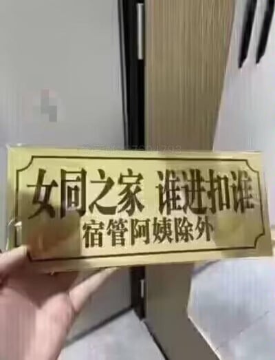 一张宿舍门牌的实拍照片，金色牌匾上用黑色字体写着“女同之家 谁进扣谁 宿管阿姨除外”的幽默标语。图片中能看到一只手正拿着这个门牌，背景似乎是宿舍走廊的环境。这种带有调侃性质的门牌常见于大学宿舍文化中，通过夸张的“谁进扣谁”表述营造轻松搞笑的氛围，同时特别注明“宿管阿姨除外”增加了现实感和趣味性。