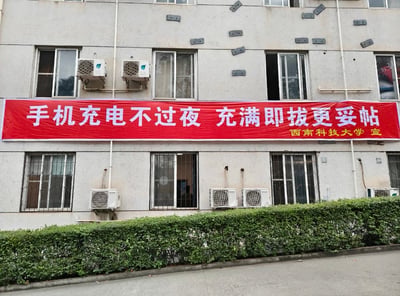 这是一张实拍的西南科技大学校园宣传照片，画面中一栋装有多台空调外机的灰白色校园建筑墙面上，悬挂着一条醒目的红色横幅。横幅以白色大字写着安全充电标语“手机充电不过夜 充满即拔更妥帖”，右下角用黄色字体标注“西南科技大学 宣”，意在提醒师生规范手机充电习惯，防范用电安全隐患，整体构图清晰，传递出明确的安全提示信息。