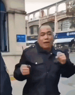 这是一段动态GIF，画面中一位穿着类似安保制服的中年大叔站在户外临街建筑旁，双手握拳抬起，做出摆动身体的搞怪动作，面部表情生动滑稽，整体氛围十分搞笑，没有双关、谐音梗或暗示内容。