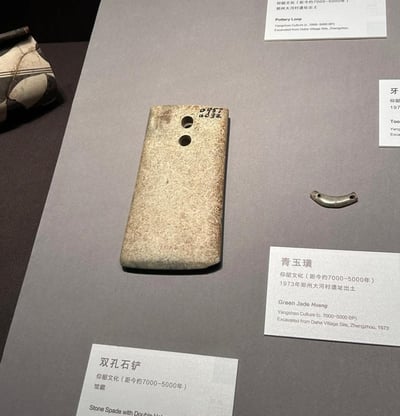这是博物馆展陈的仰韶文化文物实拍图，画面主体是一件带有双孔的石铲，表面有使用痕迹和编号，旁边搭配一件小巧的青玉璜，同时展台上还有其他仰韶文化文物的局部。每件文物都配有中英文说明牌，标注了文物名称、所属文化（距今约7000-5000年的仰韶文化）、出土地点（郑州大河村遗址）等信息，清晰展现了史前时期的生产工具与配饰器具的工艺特征，让观众直观了解仰韶文化的物质文化面貌。