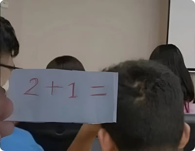 图片展示了一个课堂场景，前景中有人手持一张写有红色笔迹“2 + 1 = ”的白纸，算式结果未填写。背景中可以看到几个孩子的头顶，推测是在小学课堂上进行数学互动或提问环节，氛围轻松日常，可能用于记录教学瞬间或分享教育趣事。