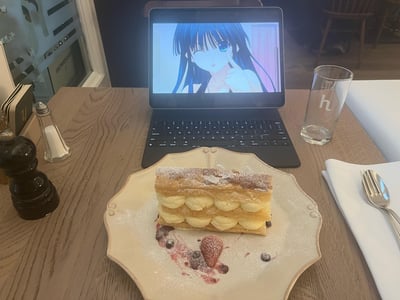 一张在咖啡厅拍摄的照片，展示了温馨的用餐场景。木质餐桌上摆放着一个精致的奶油甜点，表面撒有糖粉，点缀着草莓和蓝莓。甜点后方是一台打开的笔记本电脑，屏幕上正播放着一位蓝发蓝眼的动画少女，她正单手托腮，温柔凝视前方。桌子右侧有一个印有'h'字样的玻璃杯，旁边摆放着餐具和盐瓶、胡椒研磨器。整体画面营造出一种将二次元爱好融入现实生活的惬意氛围，展现了动画文化与美食享受的结合。