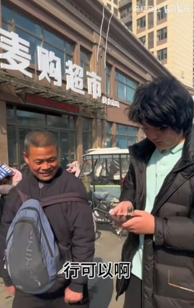 这是一张竖屏视频截图，画面展现了户外街道的日常场景：背景是带有白色大字招牌的“麦购超市”门店，一位背着蓝灰相间背包的中年大叔站在画面左侧，脸上带着憨厚的笑容，看起来心情愉悦；画面右侧的年轻男子穿着黑色厚羽绒服，内搭浅绿色上衣，正低头专注地看着手中的手机，似乎在和大叔进行沟通。画面底部配有白色字幕“行可以啊”，传递出一种轻松愉快的互动氛围，像是街头采访、随机挑战类的视频片段。