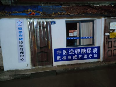 这是一张夜晚拍摄的社区医疗门店实拍图，门店为聚福唐威控糖管理中心，地址标注为营房一村40号。白色外墙的门脸处，左侧竖排标注了中心名称，右侧墙面上有醒目的蓝色大字宣传语“中医逆转糖尿病 聚福唐威五维疗法”，窗户玻璃上也印有“专注糖尿病25年 控制远离并发症”的字样。门口装有透明门帘，屋顶堆积着落叶，还有蓝色遮盖物，整体呈现出社区里小型医疗服务点的平实样貌。