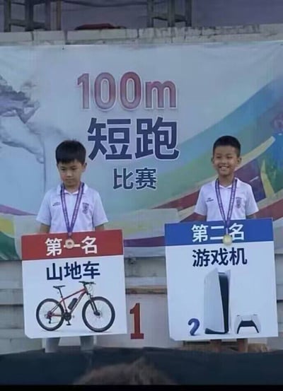 这是一张100米短跑比赛领奖场景的实拍图，背景印有“100m 短跑比赛”的字样，左侧是标注数字1的第一名领奖位，站在该位置的小男孩挂着铜色奖牌，面前标识牌显示他的奖品是山地车，他神情低落闷闷不乐；右侧是标注数字2的第二名领奖位，站在该位置的小男孩挂着金色奖牌，面前标识牌显示他的奖品是热门的游戏机，他笑容灿烂十分开心，二者的表情、获得的奖励形成强烈反差，刚好呼应标题“得了第一名为什么不开心呢”的疑问，原因是对小孩而言，第二名的游戏机奖品吸引力远高于第一名的山地车，甚至奖牌等级也更高。