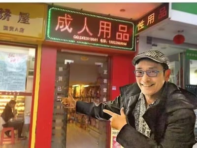 这是一张网友制作的合成P图，将演员江华的形象合成在一家成人用品店门前。江华戴着帽子和眼镜，穿着黑色外套，脸上笑容满面，双手做出指向的动作，指向身后的店铺。店铺的招牌亮着霓虹灯光，写着“成人用品”字样，门口玻璃上标注着“新货已到”“正在营业”，旁边还有“饼屋”“性用品”的店铺标识，整体画面通过这种错位合成，制造出滑稽搞笑的效果，仿佛江华在热情推荐这家成人用品店。