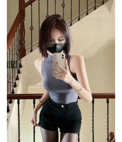 一位年轻女性在室内楼梯处自拍。她留着带有紫色挑染的短发，戴着黑色口罩和透明眼镜，身穿灰色无袖高领上衣和黑色短裤，搭配黑色丝袜。她右手拿着一部带有蛇皮纹手机壳的白色智能手机，左手自然下垂，站在楼梯扶手旁，背景是带有雕花金属栏杆的木质楼梯。