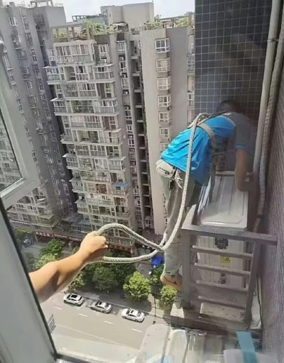 这张图拍摄于高层住宅的窗边，画面里一名穿着蓝上衣的空调安装师傅身处在室外狭窄的外机平台上，脚下是几十米高的城市街道，他身上的安全绳仅仅由室内的一只手牵拉着，安全措施极为简陋，场景惊险，让人不禁为他的安全捏一把汗，直观展现了高空作业从业者的危险工作环境与不易。