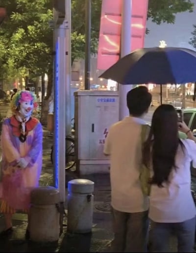 夜晚的街道旁，一位身着彩色小丑服装（头戴五彩假发、画着红鼻子和笑脸妆容，穿着红黄相间的上衣和紫色裤子）的人双手交叠站在人行道上。旁边一对情侣共撑一把深蓝色雨伞，女生正举着手机对着小丑拍照，男生侧身看向小丑。背景有树木、街灯、一个标有“公共设施”和闪电图案的配电箱，以及亮着粉色灯光的大型广告牌，整体呈现出一种城市夜晚街头的日常又略带趣味的场景。