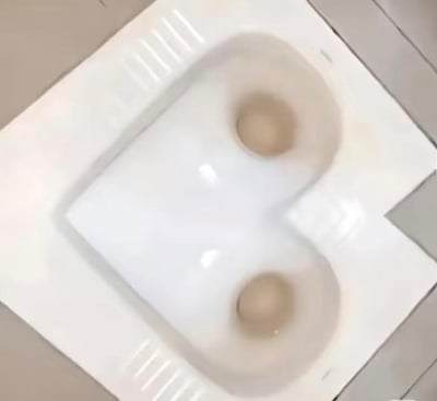 这是一张实拍的白色蹲便器照片，蹲便器的整体凹陷轮廓加上两个圆形的下水孔，在视觉上恰好构成了一张人脸的模样，两个下水孔如同人脸的眼睛，造型十分滑稽搞笑，给普通的卫浴设施赋予了趣味的视觉效果，没有双关、谐音梗等特殊含义。