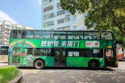 一辆绿色双层巴士停在城市街道上，车身印有大型青岛啤酒广告，广告标语为“想炉管 来厦门”，旁边有两瓶绿色啤酒瓶图案。背景是蓝天白云下的居民楼和绿树，画面清晰明亮。

**谐音梗说明**：标语“想炉管”可能是“想旅游”的谐音误用或方言表达，结合“来厦门”形成疑似旅游宣传的歧义文案，因“炉管”在普通话中意为烟囱，导致语句不通顺而产生搞笑效果。